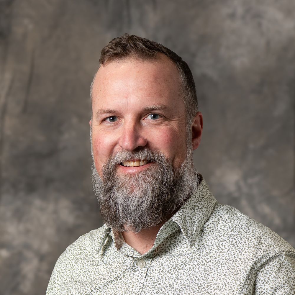Portrait of Stefan Wild, smiling and wearing a patterned shirt, set against a neutral gray backdrop.