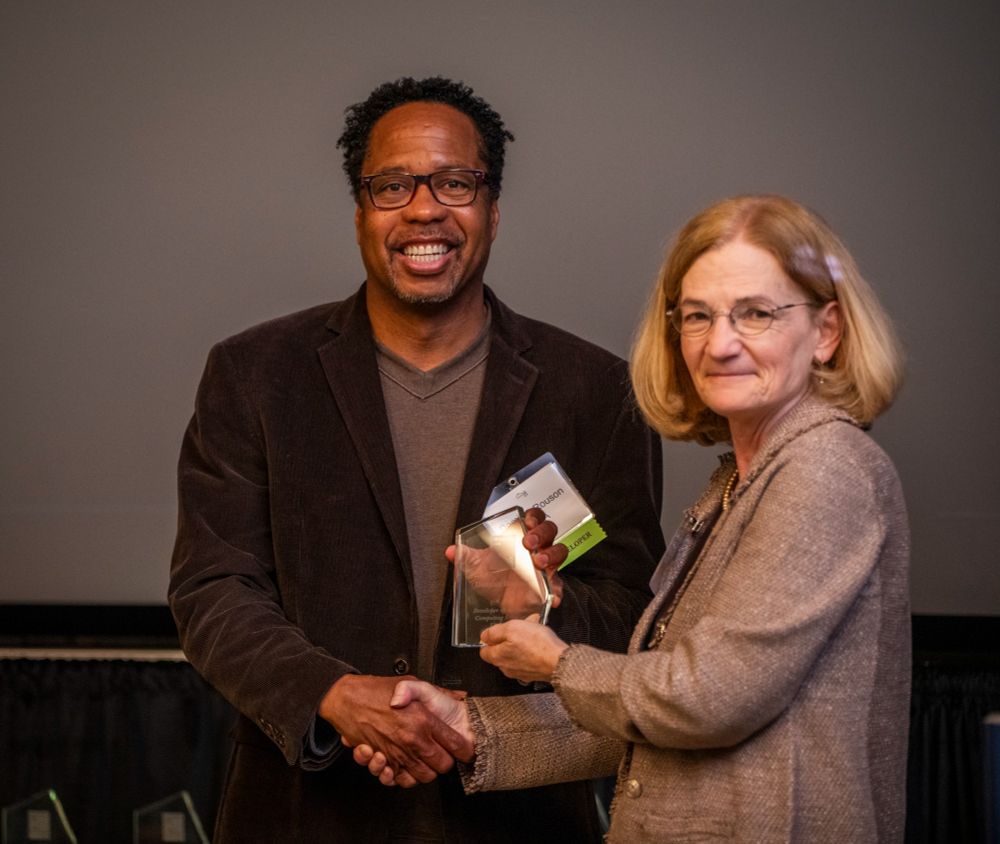 Two individuals are posing for a photo at an event. On the left, a smiling man in a brown blazer and glasses is holding an award. On the right, a woman in a beige suit and glasses is shaking his hand while also smiling slightly. Both appear to be in a formal setting with a plain backdrop.