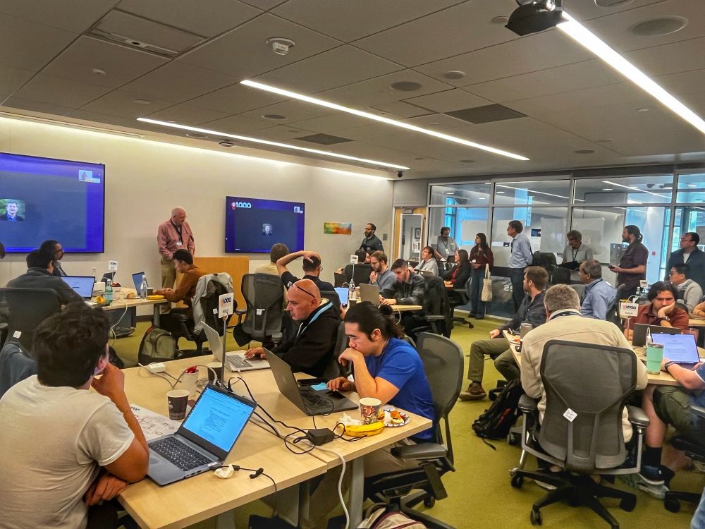 A room full of scientists and researchers participate in a collaborative computing event at Berkeley Lab. Attendees sit at tables working on laptops, engaging in discussions, and taking notes. A speaker at the front of the room presents at a podium while two large screens display virtual participants. Others stand along the walls, observing and interacting. The setting is a modern workspace with glass walls and bright lighting.