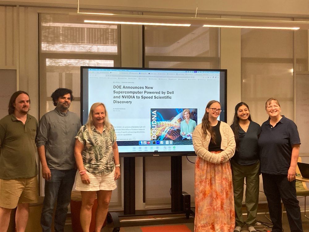Six people stand in a row in front of a large screen displaying a news article titled "DOE Announces New Supercomputer Powered by Dell and NVIDIA to Speed Scientific Discovery." The setting appears to be a modern conference or classroom space with glass windows and bright lighting.