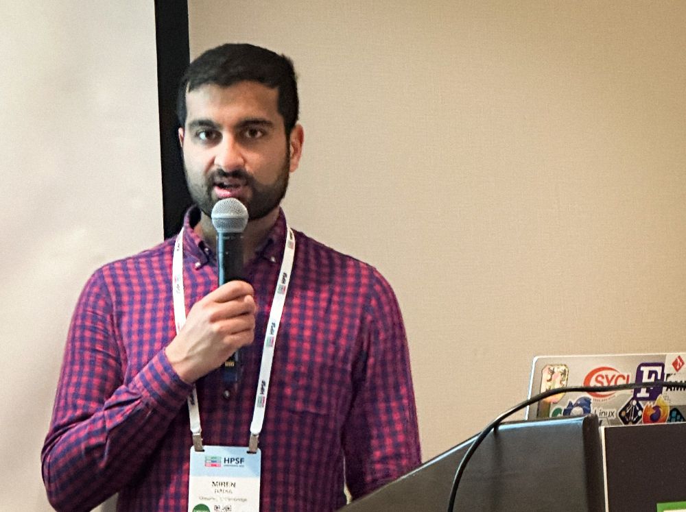 Person wearing a red and blue plaid shirt stands at a podium holding a microphone, with a conference badge and lanyard visible. A laptop covered in stickers sits on the podium.