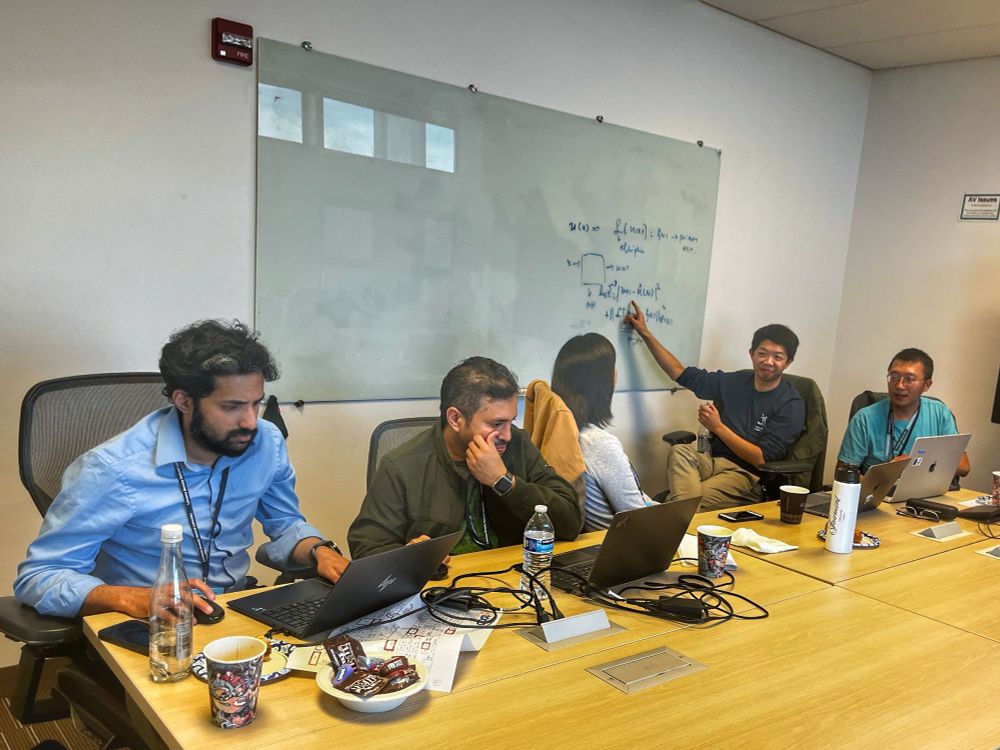 A group of five people is gathered around a conference table in a meeting room. One person is writing on a whiteboard, explaining a concept to the others. The whiteboard contains mathematical equations and diagrams. Three participants are working on laptops, while two are engaged in discussion. The table has coffee cups, water bottles, and snacks. The setting appears to be a collaborative work session, possibly related to scientific or technical research.