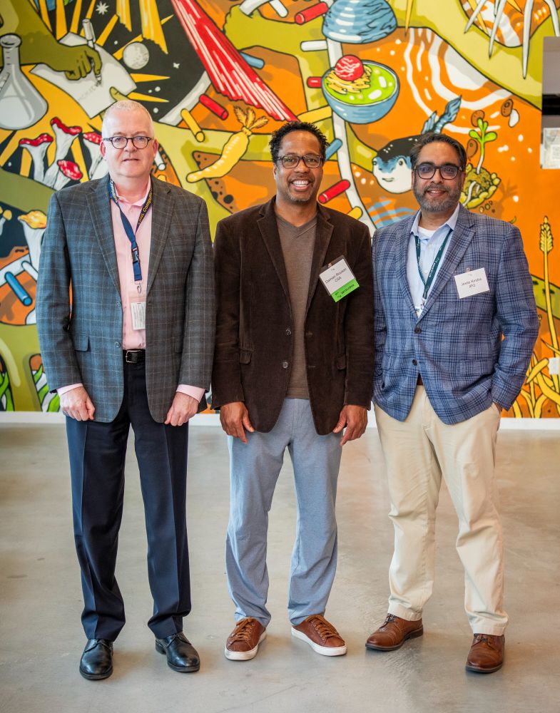 Three men are standing side by side, smiling for a photo in front of a colorful, science-themed mural. The man on the left is wearing a plaid blazer and glasses, the man in the center is in a brown blazer and casual trousers, and the man on the right is in a blue plaid blazer and khaki pants. The setting appears to be a professional or academic event.