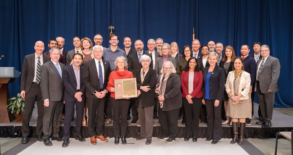 People standing on stage in front of a blue curtain accepting an award from the Secretary of Energy, Jennifer Granholm.