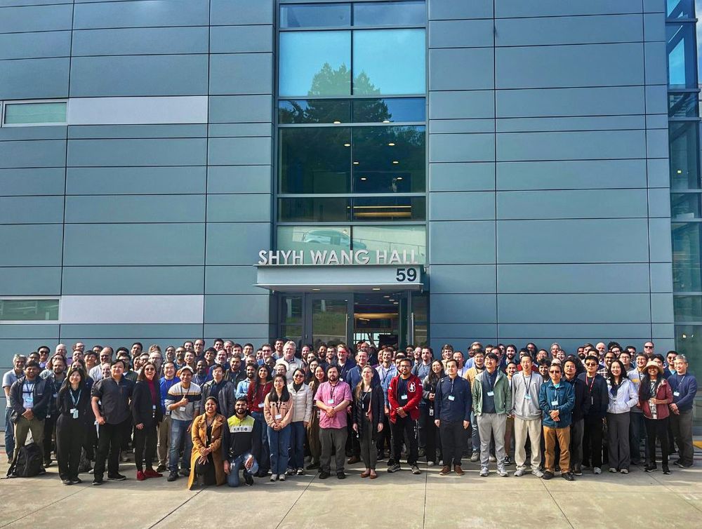 A large group of scientists and researchers gather for a group photo in front of Shyh Wang Hall at Berkeley Lab. The participants, diverse in age and background, stand together on a sunny day, smiling at the camera. The modern glass and metal facade of the building serves as the backdrop, with the building name and number "59" visible above the entrance.