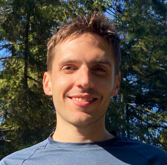 A person standing outdoors in front of tall green trees, wearing a dark crew-neck shirt in natural sunlight.