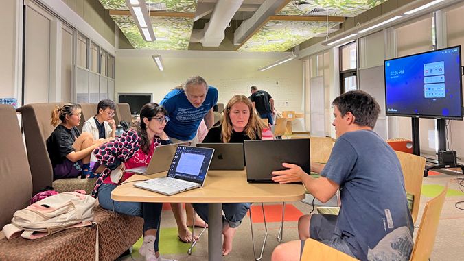 A group of people are gathered around a table in a modern classroom or collaborative workspace, working together on laptops. Some individuals are seated while others are standing and engaging in discussion. In the background, a large screen displays a digital interface, and the room features bright lighting and patterned ceiling panels. The atmosphere appears focused and collaborative.