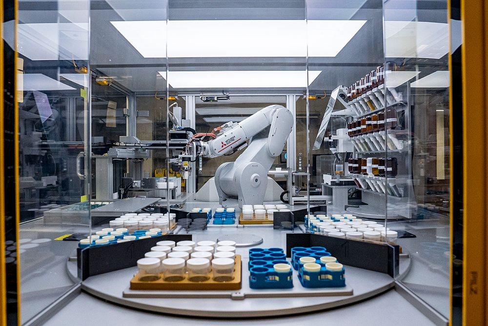 A robotic arm is positioned in a high-tech laboratory setting, surrounded by transparent glass panels. The robot is engaged in materials testing, with various containers organized on a circular platform in the foreground. On the right, shelves hold additional sample jars, highlighting the automated setup designed for efficient scientific research. Bright overhead lights illuminate the workspace.