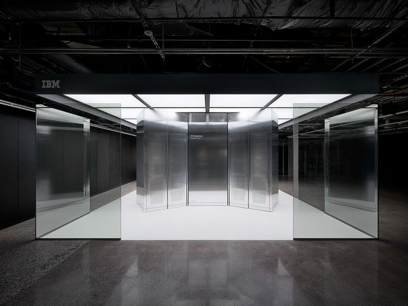A sleek, futuristic IBM quantum computer system displayed in a minimalistic, glass-enclosed environment with polished concrete floors and overhead industrial lighting. The IBM logo is visible on the upper left corner of the enclosure.
