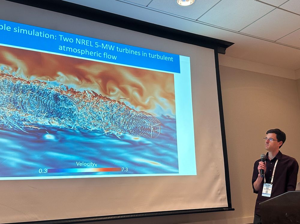Presenter stands at a podium with a microphone in front of a projection screen displaying a simulation visualization of two wind turbines in turbulent atmospheric flow, with a color bar indicating velocity.