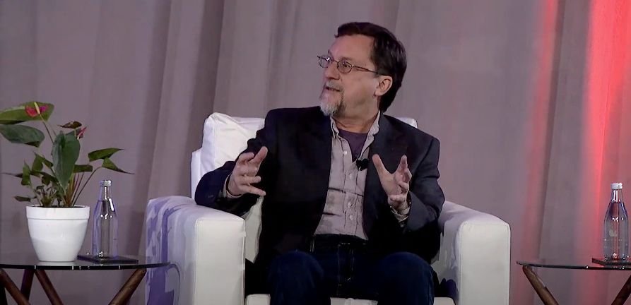 A man with glasses and a beard, wearing a dark blazer and a button-up shirt, sits on a white armchair while speaking and gesturing with both hands. He appears engaged in a discussion on stage, with a gray curtain backdrop and red accent lighting. A small table with a potted plant and water bottles is visible beside him.