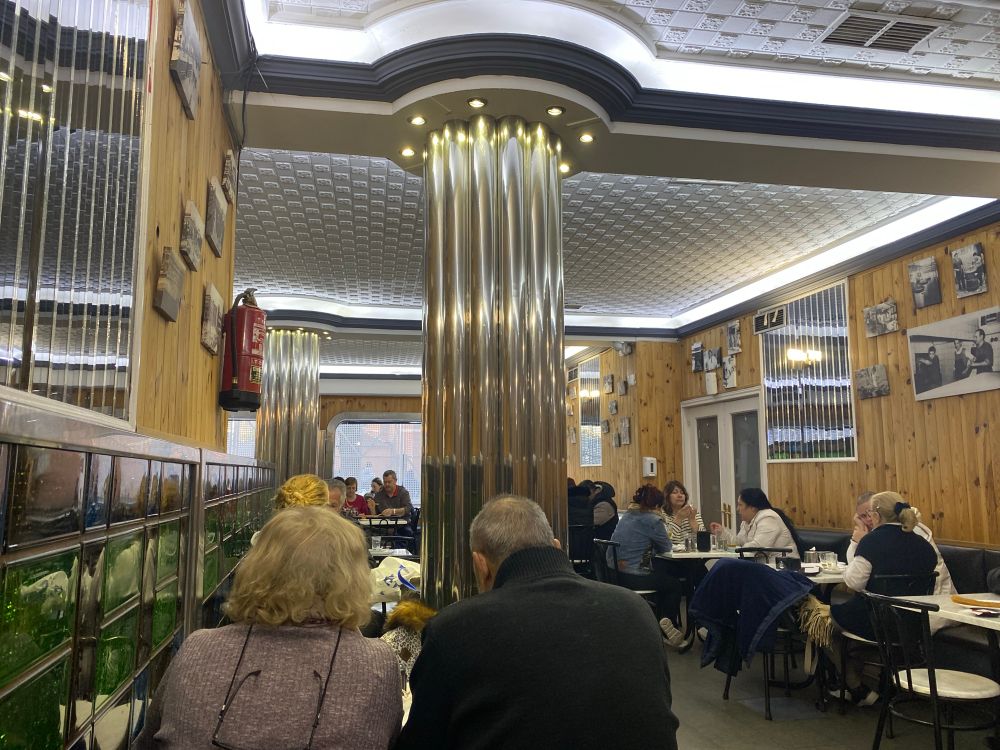Interior de una cafetería de estilo antiguo, con superficies de metal, madera y vidrio. Personas desayunando y en primer plano una mujer y un hombre mayores.