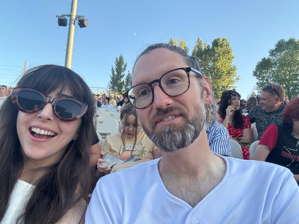 White woman and white man selfie at an outdoor concert venue. She is smiling with oversized glasses, he is maybe about to say something but just looking goofy