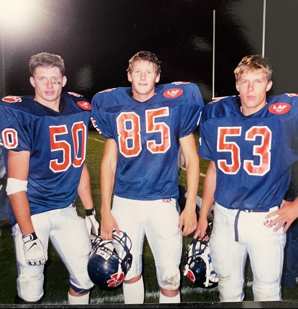 Three high school kids in football uniforms: navy blue jerseys with orange numbers and white pants