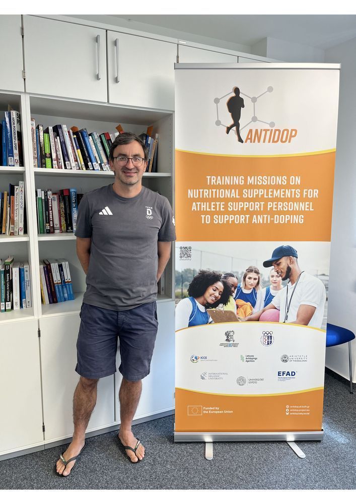 Dr. Christian Heiss standing next to the AntiDop roll-up banner after delivering a guest lecture at Leipzig University. The banner shows the project logo, title, a thematic image, and the partner institution logos.