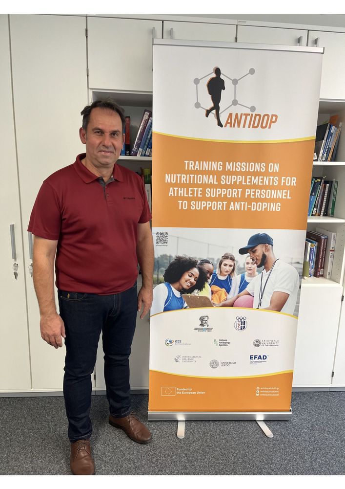 Ioannis Morres from the University of Thessaly standing next to the new AntiDop roll-up banner during his visit to Leipzig University. The banner displays the project logo, title, a thematic image, and the logos of all partner institutions.