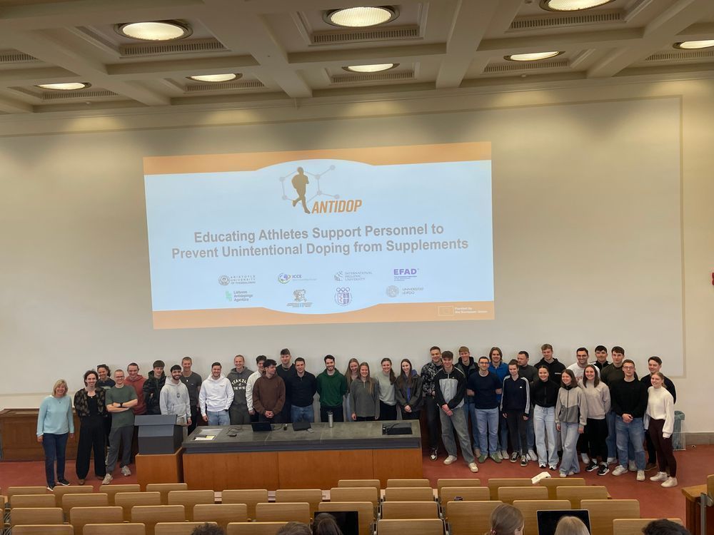 Prof. Dr. Anne-Marie Elbe and a group of university students smiling and posing together in front of a projected AntiDop project slide in a lecture hall at Leipzig University.