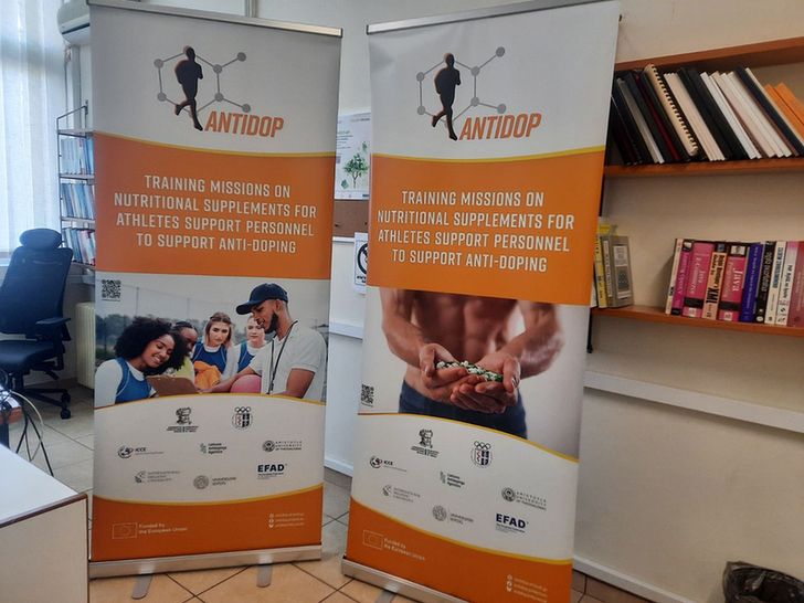 Front view of the AntiDop project roll-up banner showing the logo, project theme, a visual related to clean sport, and logos of all partner institutions. The banner is standing indoors, ready for use at events.