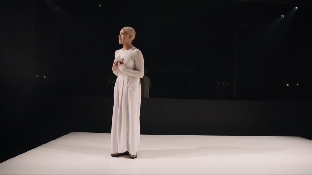 Cush Jumbo stands dressed in white, on a white stage, grasping a white paper to her chest