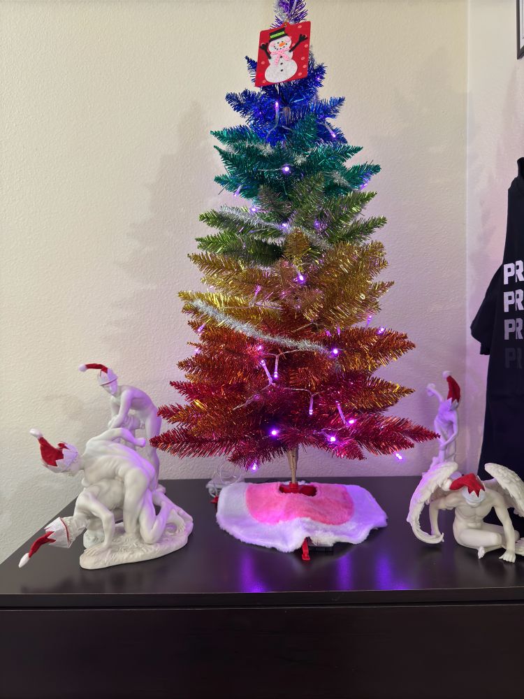 A tabletop Pride rainbow colored Christmas tree surrounded by 4 small nude Greek statues wearing Santa hats.