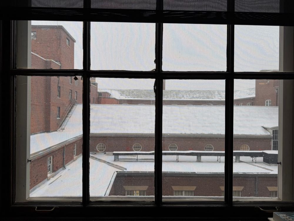 View out of a window as it lightly snows, capturing the building’s slanted roofs and windows. 