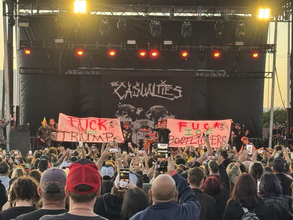 Outdoor punk concert at dusk. Crowd of people hold up smartphones toward a lit stage where a band performs beneath a large “The Casualties” backdrop. Two hand-painted banners onstage display: “FUCK YOU TRUMP” and “FUCK ICE BOOTLICKERS.” Lights glow red and yellow.