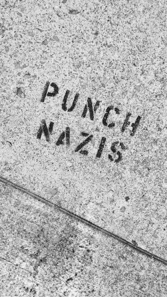 A rough spray-paint stencil on a light-gray concrete sidewalk reads PUNCH NAZIS in bold, uppercase black letters. The two words are stacked: “PUNCH” on top of “NAZIS” that sit at a slight diagonal, about 20° tilted counterclockwise. The concrete slab is speckled with small stones and fine cracks; a seam line cuts diagonally across the lower third of the image.