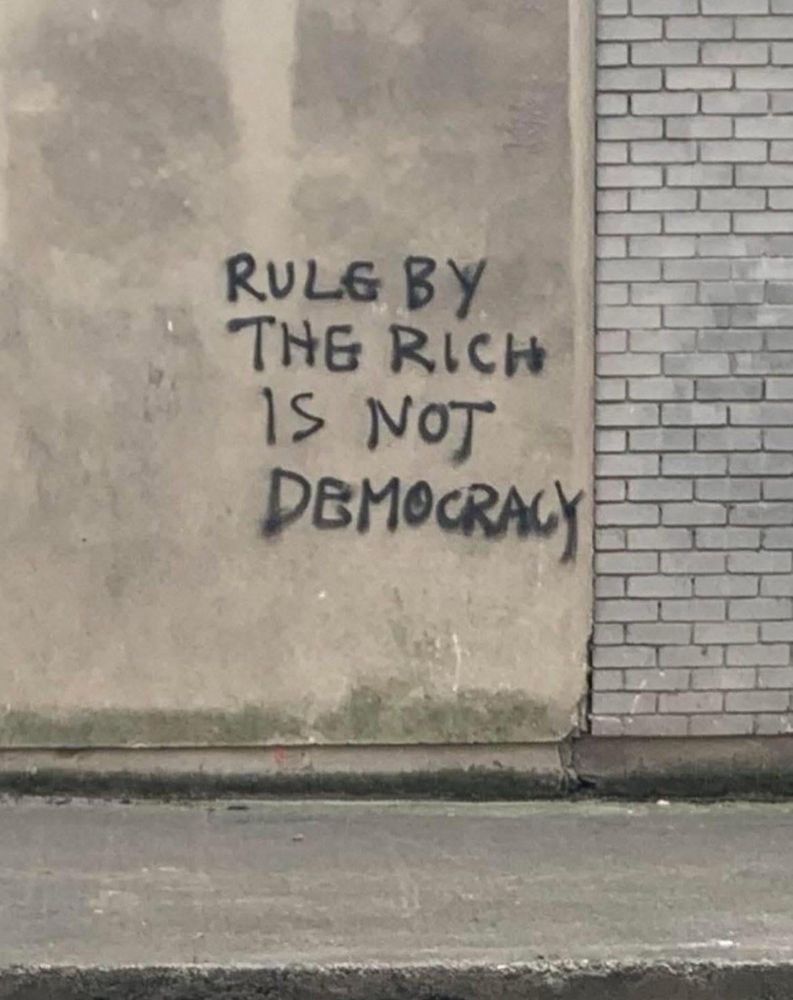 The image shows graffiti spray-painted in black on a worn, beige concrete wall next to a section of light gray brick. The text reads in all capital letters:

“RULE BY
THE RICH
IS NOT
DEMOCRACY”

The letters are uneven and hand-painted, giving it a rough, urgent appearance. The wall has visible stains and weathering, and the ground in front of it is a gray, wet-looking pavement.