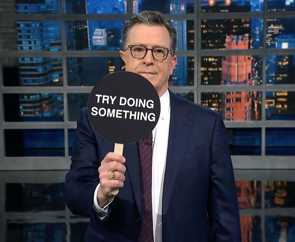 The image shows Stephen Colbert wearing a formal navy-blue suit and glasses, standing in front of a nighttime cityscape backdrop typical of his late-night talk show set. He holds a round black sign on a wooden stick, prominently displaying bold white text that reads, “TRY DOING SOMETHING.” His neutral, direct expression reinforces a clear call to action or critique, characteristic of Colbert’s humorous yet impactful style.