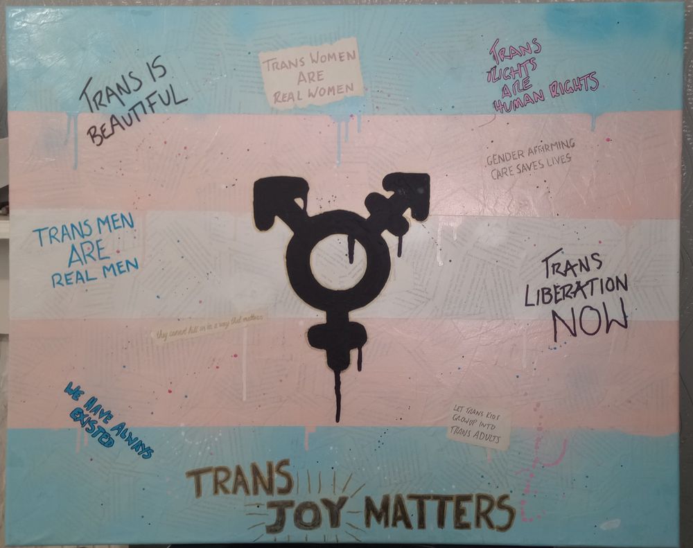 A trans positive painting on canvas.

The background is a collage of pages torn from a book with a trans pride flag painted over the top. In the centre is a large trans symbol, painted in black and outlined in gold. The paint of the flag and the symbol have been intentionally allowed to drip. Written about the painting are the following phrases:

- trans is beautiful
- trans women are real women
- trans rights are human rights
- trans men are real men
- gender affirming care saves lives
- they cannot kill us in a way that matters
- trans liberation now
- we have always existed
- let trans kids grow up into trans adults
- trans joy matters