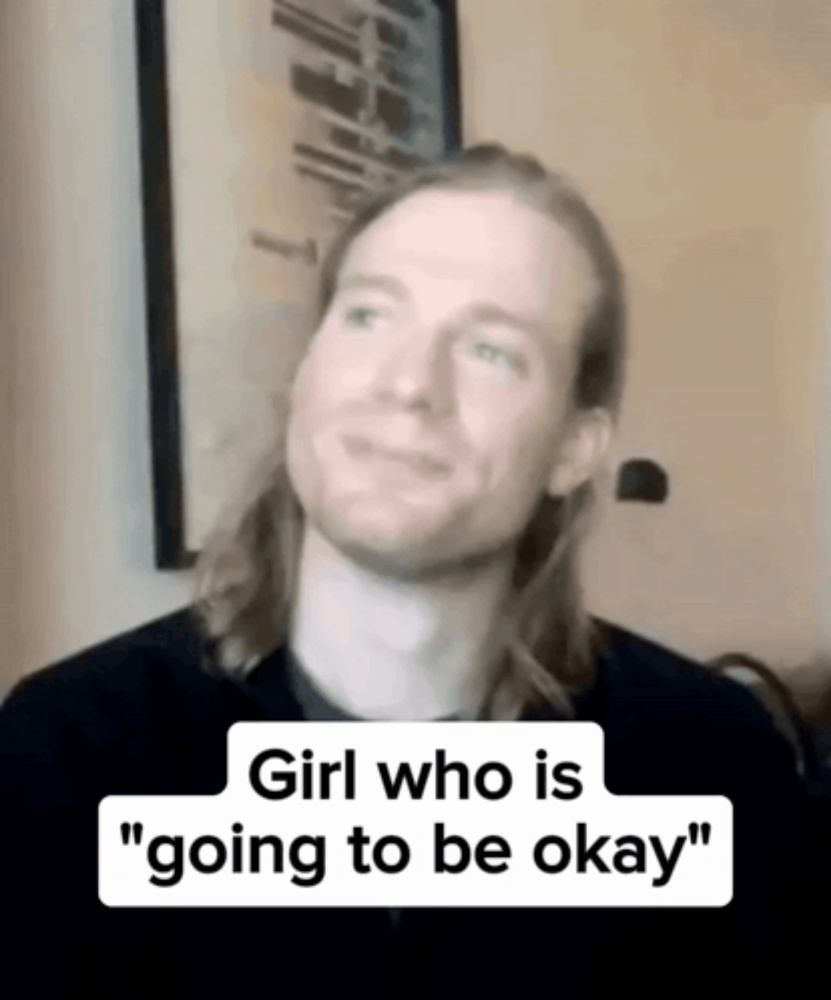 a man with long hair is sitting in front of a wall with a sign that says girl who is going to be okay .