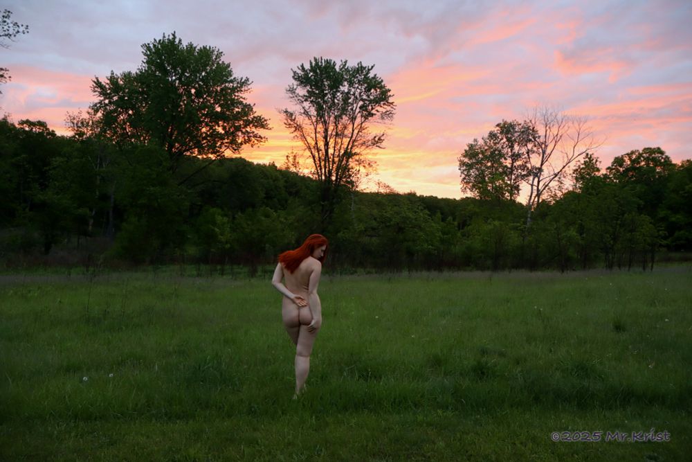 Nude female model with red hair and pale skin, seen from the back, against a green field, trees, and colorful sunrise.
