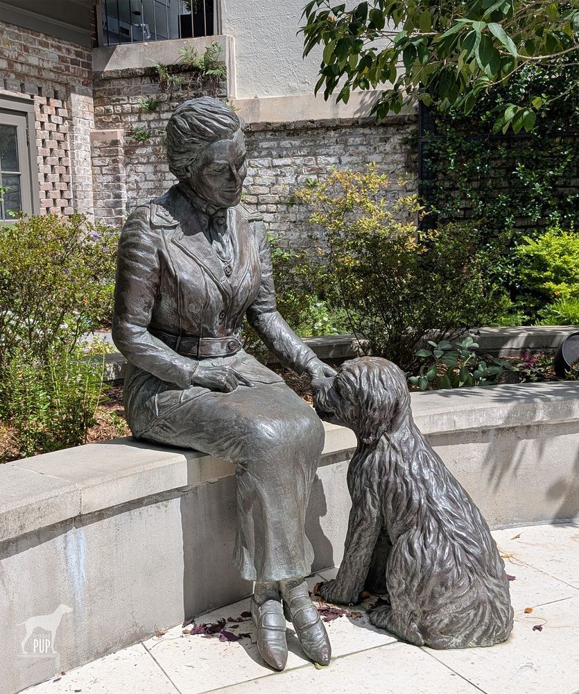 Statue pairing of a seated woman wearing a long skirt with her left arm outstretched petting a seated, mid-sized shaggy dog facing her.