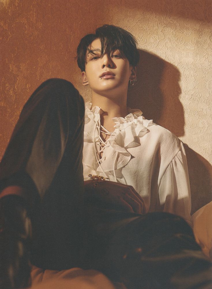 Jungkook special 8 photo folio (time difference). He is sitting, wearing a shirt with a frilly collar, and black dress pants. We can see the tip of his black boot. The light is in brown/beige tones and is projected in such a way that his shadow is drawn on the wall he is leaning against