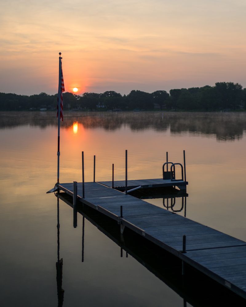 American Flag at the end of a dock with the rising sun in the background over a still lake.