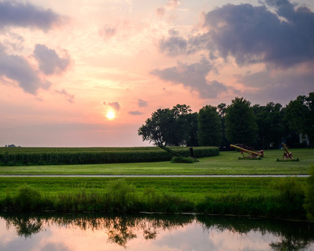 The setting sun over the fields and farms of Iowa with a pond in the foreground.