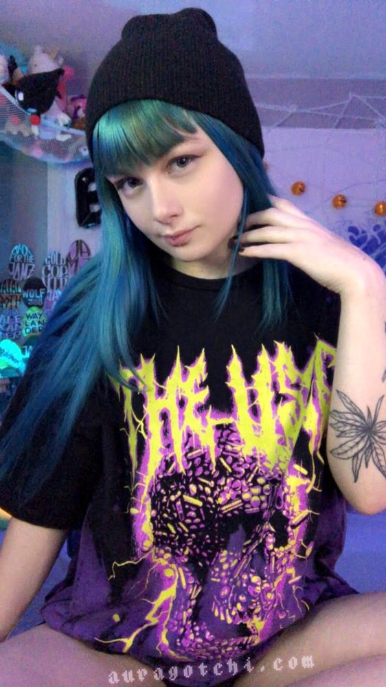A camgirl with long teal-blue hair wears a black beanie and a graphic oversized black T-shirt featuring a vivid purple and yellow design with the text "THE USED." They are sitting in a colorful room with plush toys, posters, and string lights in the background. A large cannabis tattoo is visible on their arm, and the website "auragotchi.com" is written at the bottom.