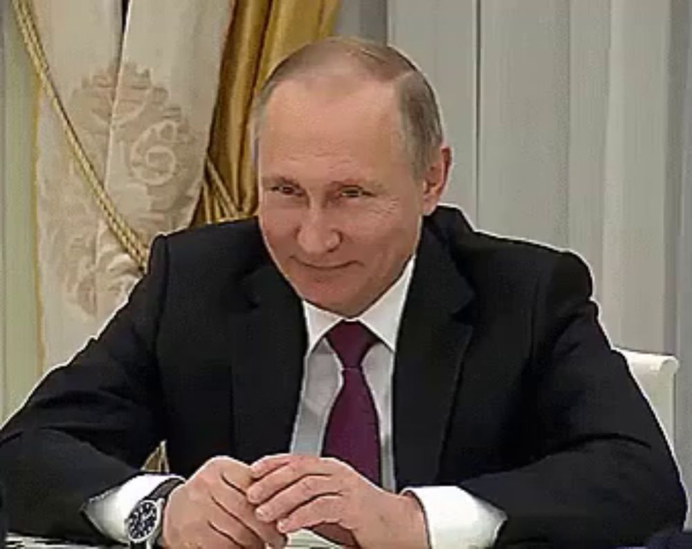 a man in a suit and tie is smiling while sitting at a table with his hands folded .