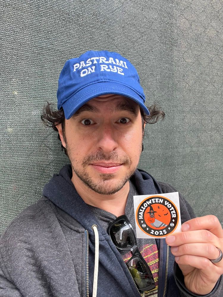 Me wearing a pastrami on rye baseball cap and holding a Halloween voter sticker 