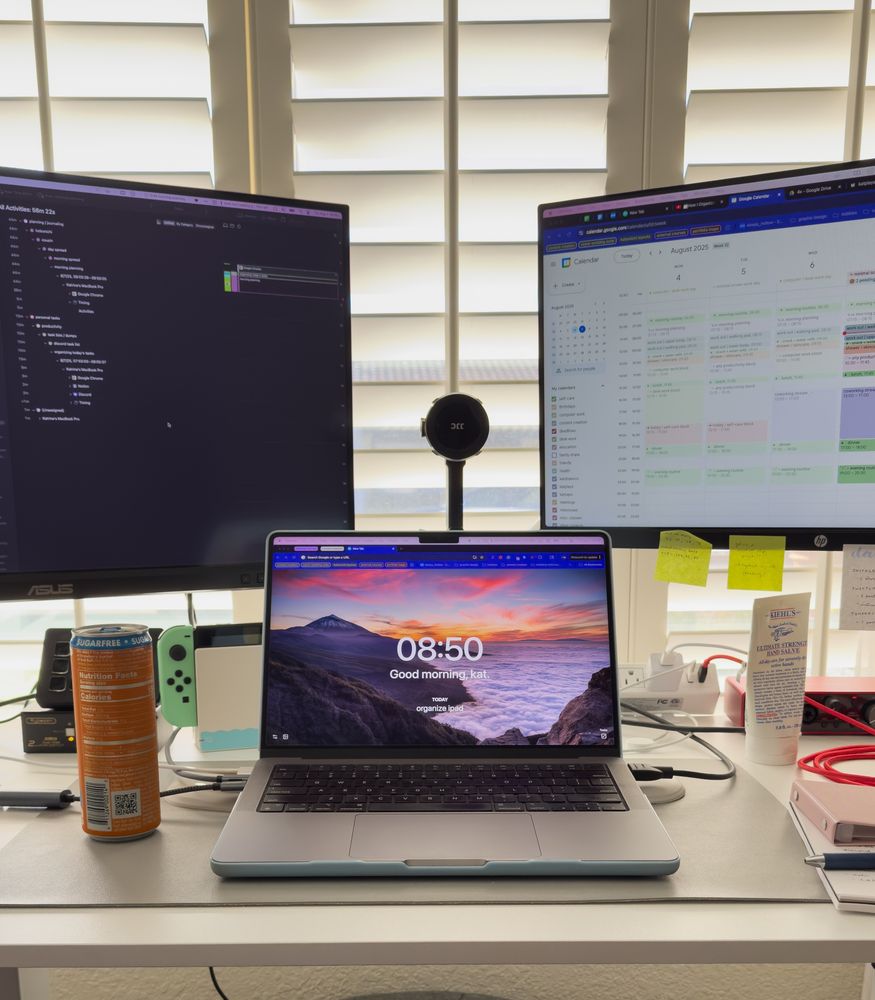a picture of a desk with a laptop and two monitors behind it to its left and right. next to the laptop sits an orange energy drink can on the left and to the right sits kiehls hand cream. behind the laptop is a nintendo switch and on the desk are various other tech devices and stationery supplies.