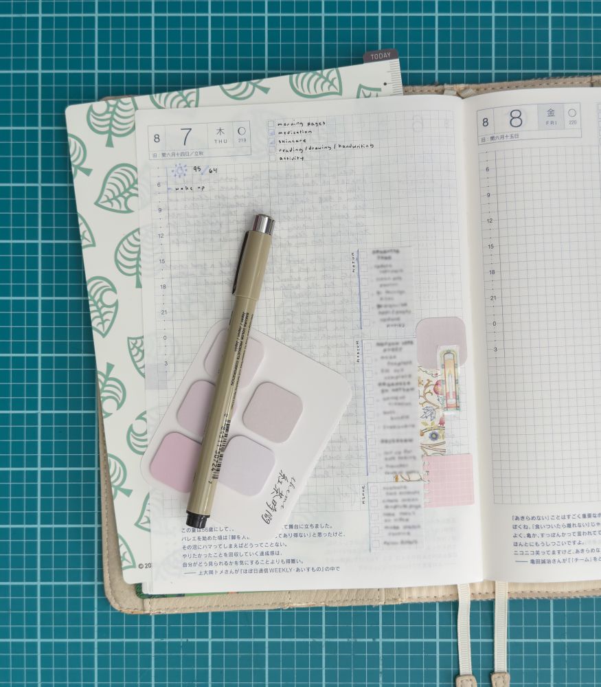 a hobonichi planner sitting on a green desk mat open to the daily page on the left labeled as august 7th. the planner has a brown cover and the page on the left has a green animal crossing pencil board underneath it. various patterned and colorful sticky notes / washi tape are pasted on the page underneath to do list sticky notes organized by category with a page of sticky notes in pastel pink and beige tones and a micron pen sitting on top.