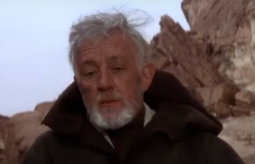 Old Ben Kenobi with gray hair and a gray beard in a hooded robe with the rocky terrain of Tatooine in the background.