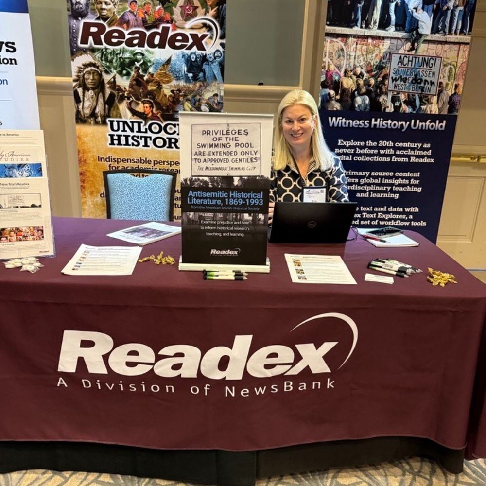Readex table at the Charleston Conference vendor showcase. Table covered in a maroon table cloth with the Readex logo on the front in white. Account representative Erin sits at the table with a laptop. Flyers are lying on the table. Two banners appear in the background. On the left the banner features a collage of historical figures with Readex superimposed over the collage. The banner on the right features an image of the fall of the Berlin Wall.