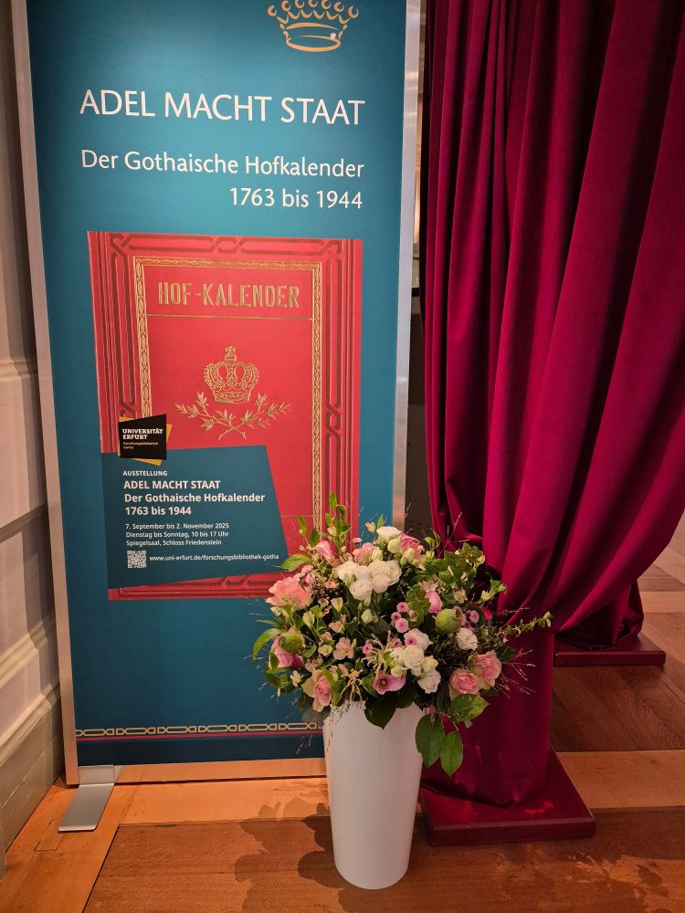 Plakat der Ausstellung am Eingang des Spiegelsaals neben einem roten Samtvorhang. Im Vordergrund steht eine weiße Vase mit Blumen auf dem Boden.