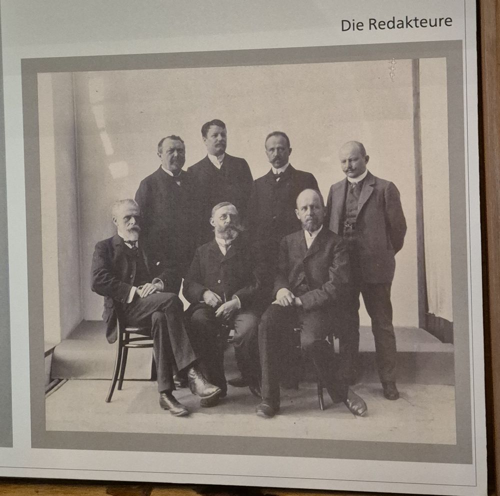 Ein Foto in der Ausstellung zeigt die Redakteure des Gothaischen Hofkalenders; drei von ihnen sitzen auf einem Stuhl, vier stehen dahinter.