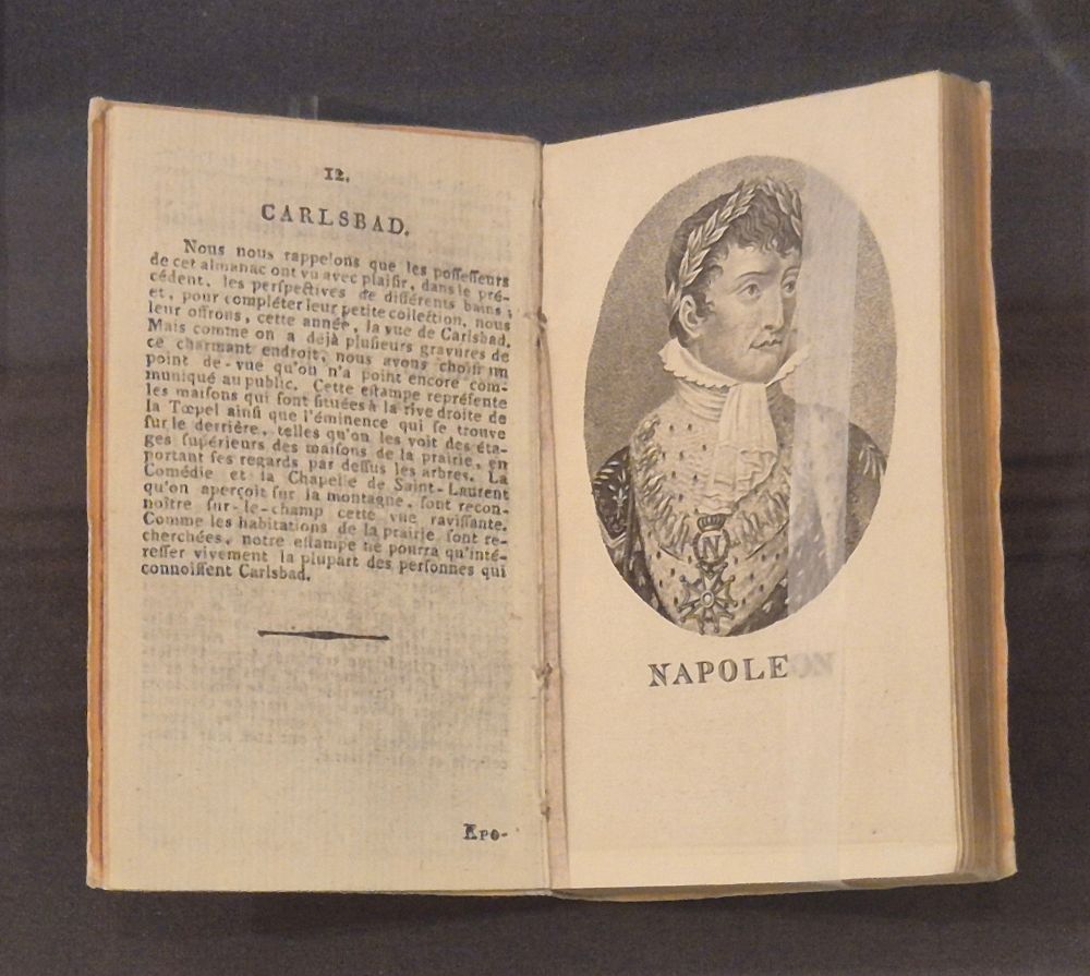 Ein aufgeschlagenes Buch. Auf der linken Seite befindet sich ein französischsprachiger Text, betitelt mit "Carlsbad". Auf der rechten Seite ist das Porträt Napoleons I. Bonaparte.