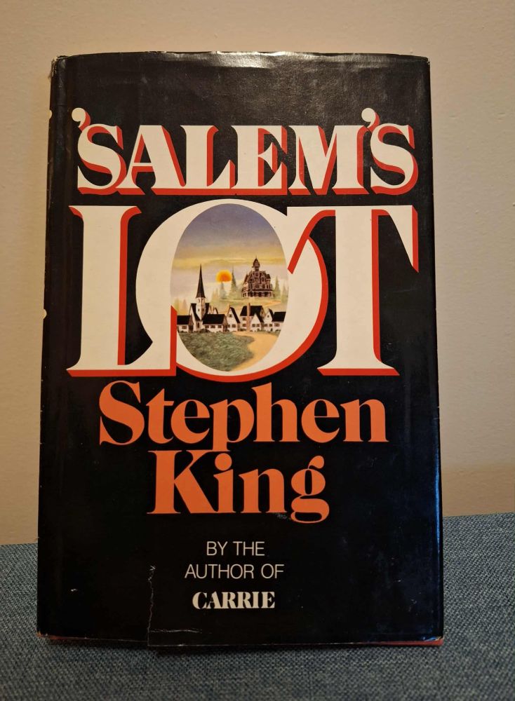 Cover of hardcover with book sleeve 'Salem's LOT - black cover with title, the 'O' has image insideof the town, church, and the Marsten House sitting high on a hill, Stephen King in orange type, below in smaller white type, BY THE AUTHOR OF CARRIE
