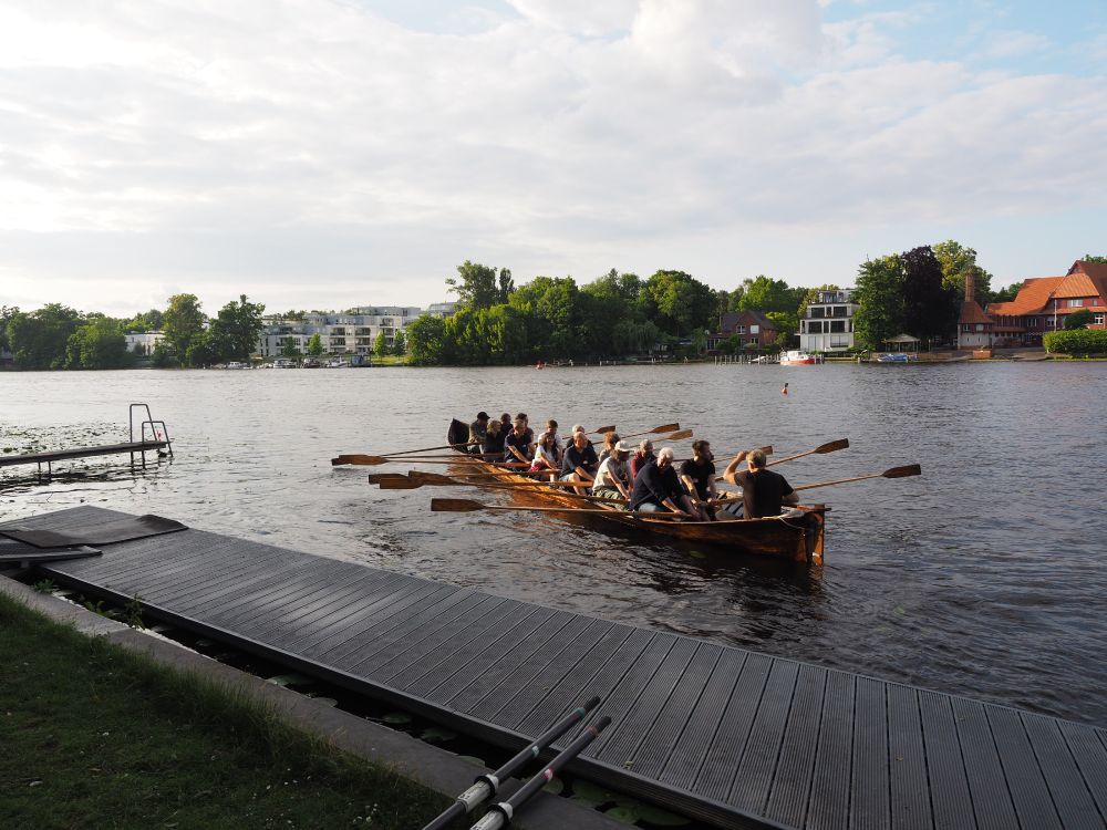 Eine Gruppe von Menschen rudert gemeinsam in einem traditionellen Holzboot auf einem ruhigen Fluss, umgeben von grünen Bäumen und Uferhäusern.