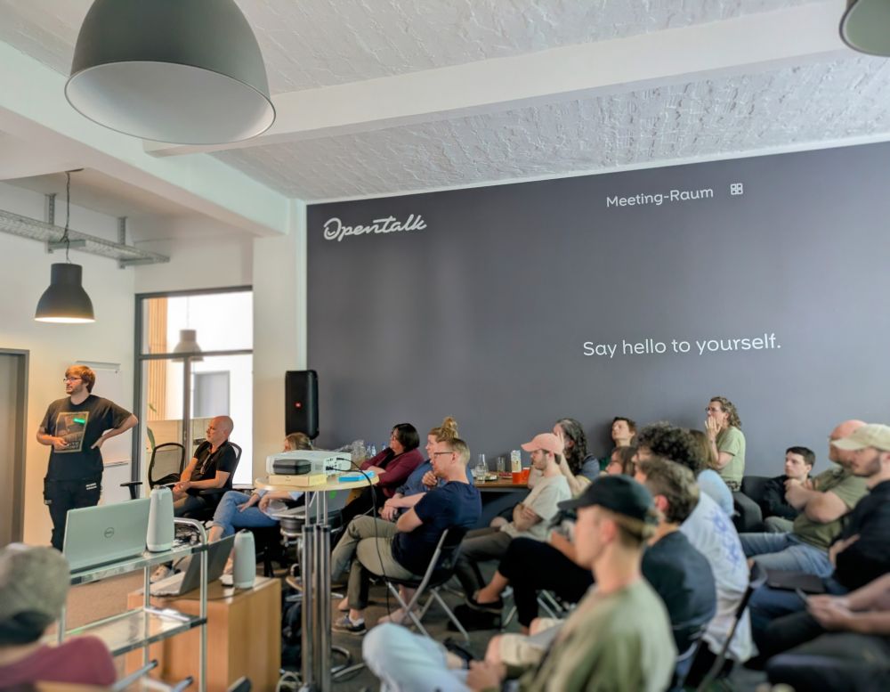 Ein Team sitzt aufmerksam in einem modernen Meetingraum von OpenTalk, während ein Kollege vorne eine Präsentation hält. An der Wand steht der Schriftzug „Say hello to yourself.“.