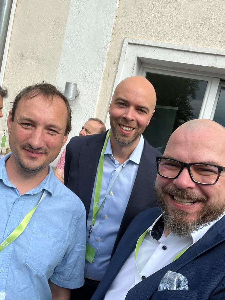 Drei Männer mit Namensschildern und grünen Lanyards lachen in die Kamera – aufgenommen bei einer Networking-Pause auf dem Summit.
